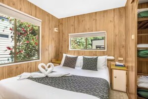 Quarto com parede de painel de madeira, cama com colcha estampada, duas almofadas, mesa de cabeceira e janela com vista para vegetação.