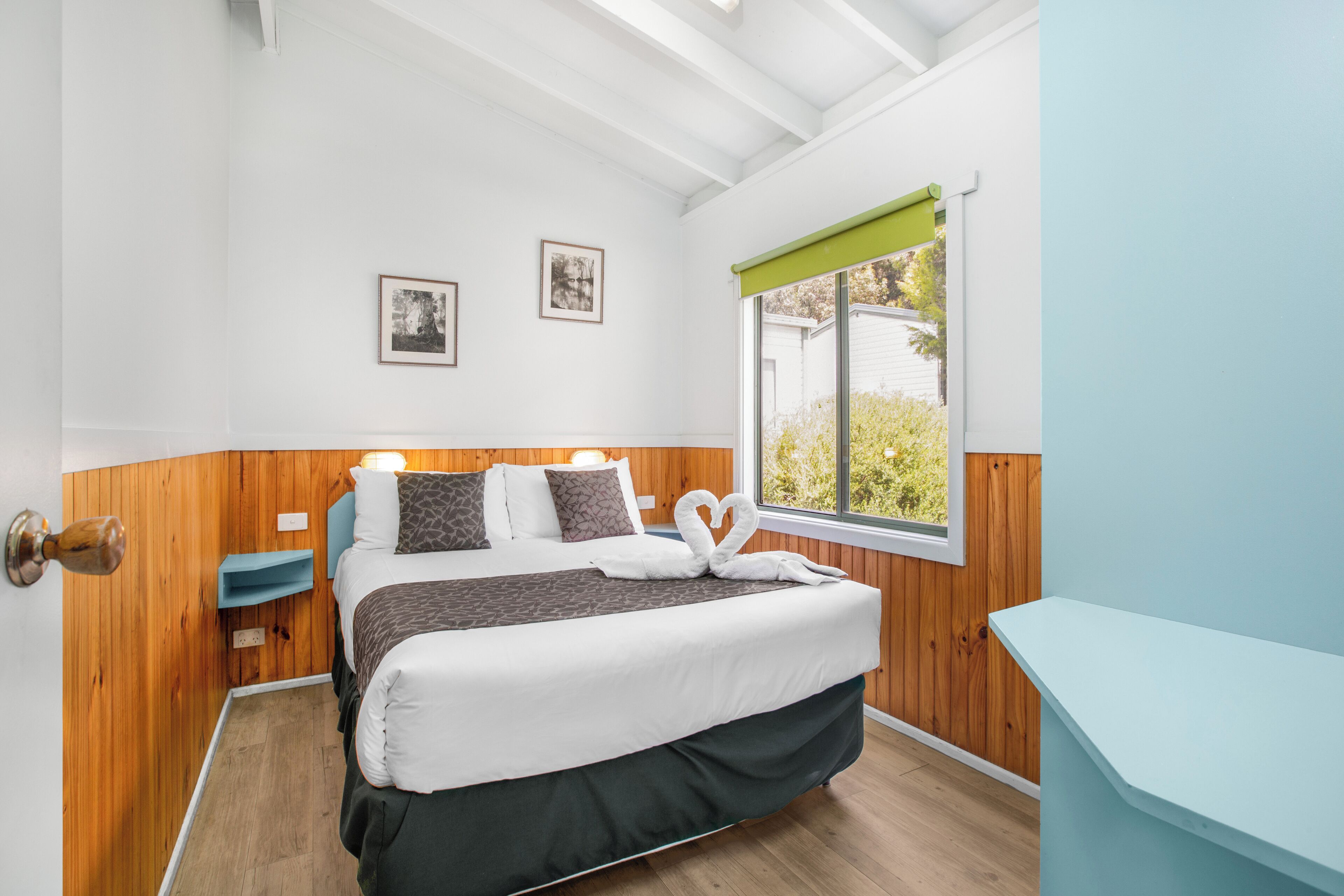 A neatly arranged bedroom with a bed, bedside tables, framed pictures on the wall, and a window with a view of greenery.