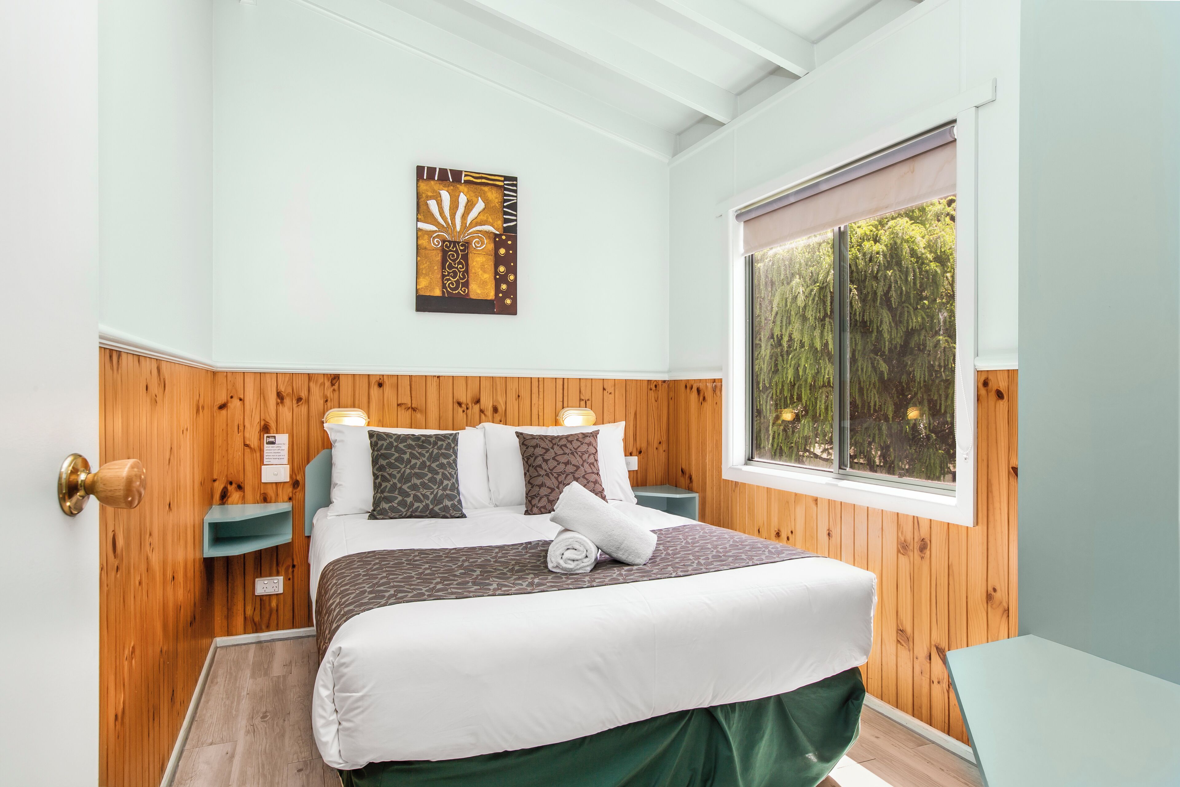 A bedroom with a wooden paneled wall, a bed with white bedding, a green skirt, a window with a view of trees, and a wall-mounted artwork.