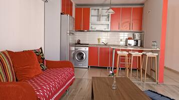 Un espacio habitable funcional con sofá, una mesa de centro de madera, lavadora y una kitchenette con armarios rojos y un microondas.