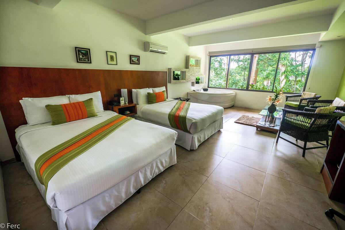 Habitación de hotel con dos camas, un escritorio, una silla y un amplio ventanal con vistas a zonas verdes.