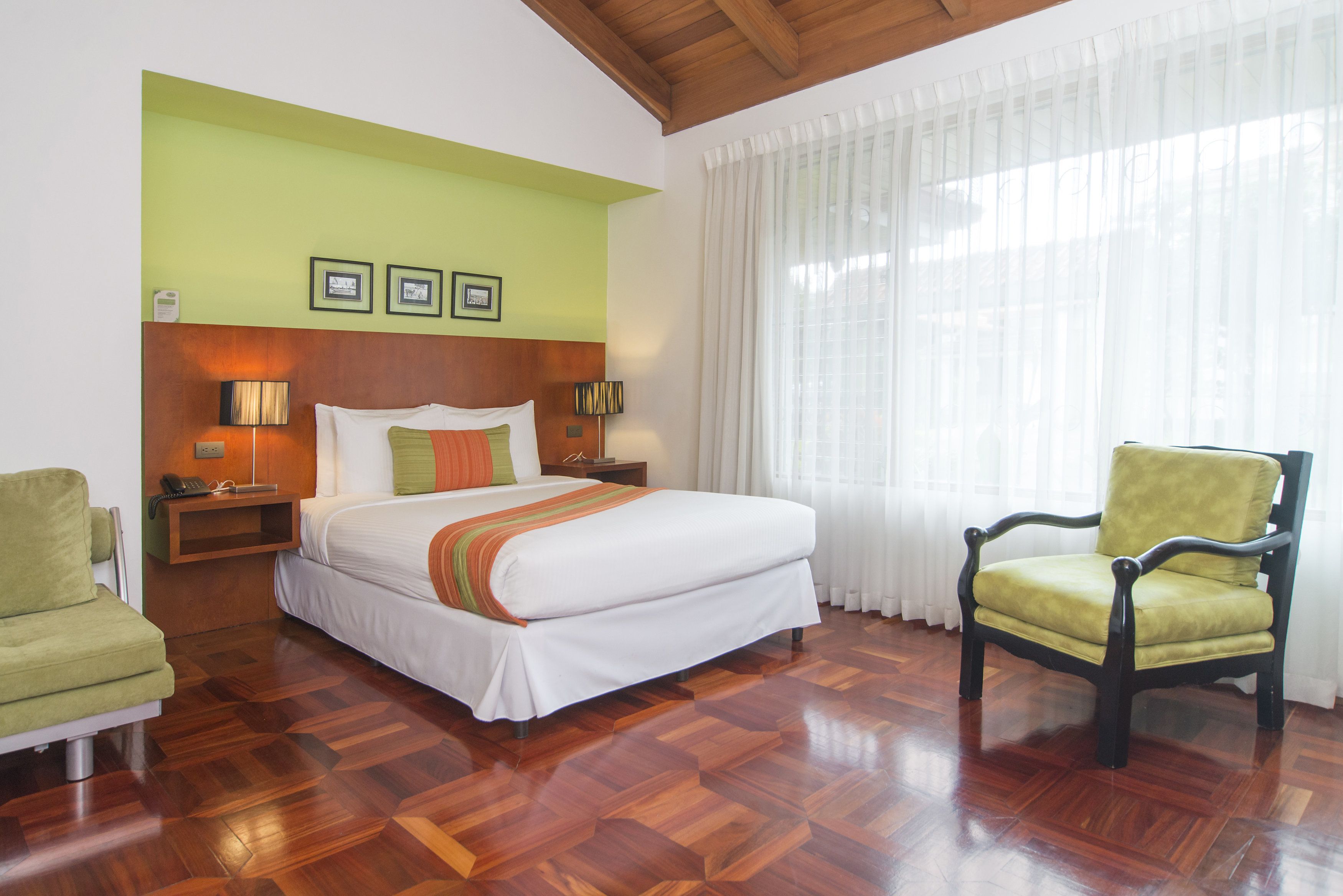 A hotel room with a large bed, a green armchair, a wooden floor, and a window with sheer curtains.
