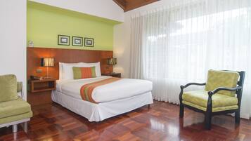A hotel room with a large bed, a green armchair, a wooden floor, and a window with sheer curtains.