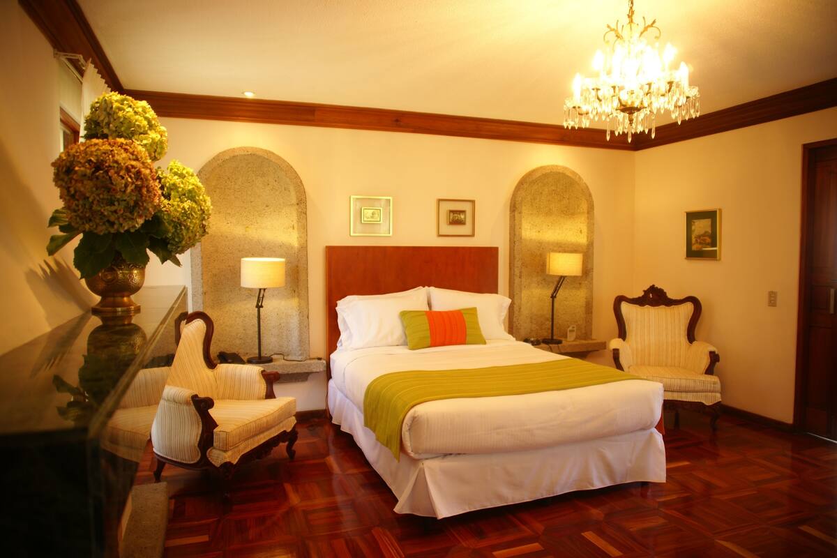 Una habitación de hotel con una cama grande, dos sillas, una araña y suelo de madera.