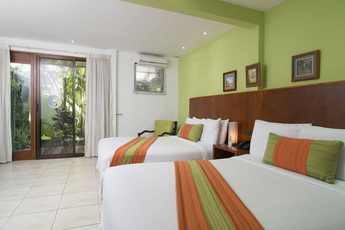 Habitación de hotel con dos camas, una puerta corrediza de vidrio a un balcón, cabecera de madera y una pequeña mesa con lámpara.