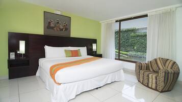 Habitación de hotel con una cama grande, una mesita de noche, una silla y una ventana con vistas a zonas verdes.