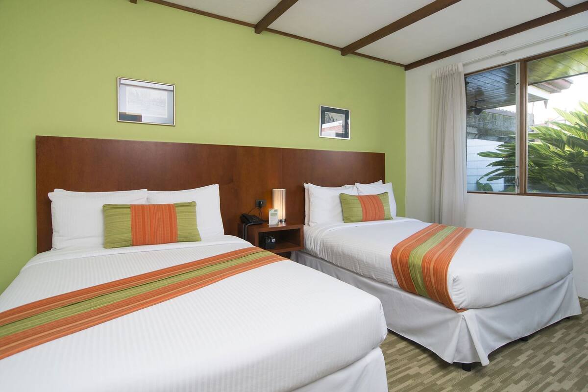 Habitación de hotel con dos camas, cabecera de madera, mesita de noche, lámpara y ventana con cortinas.