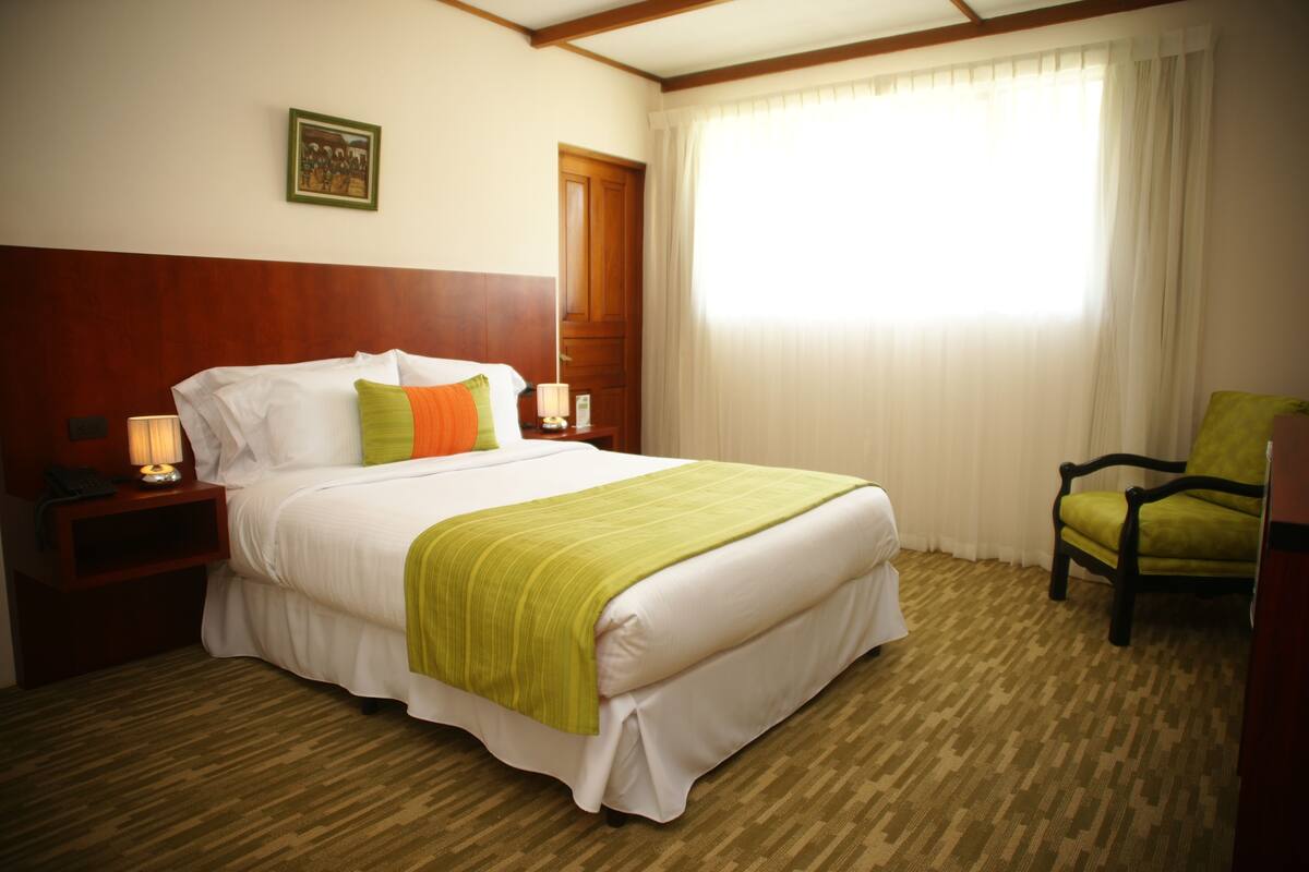 Habitación de hotel con una cama grande, cabecera de madera, manta verde, mesita de noche con lámpara, una silla y una ventana con cortinas.