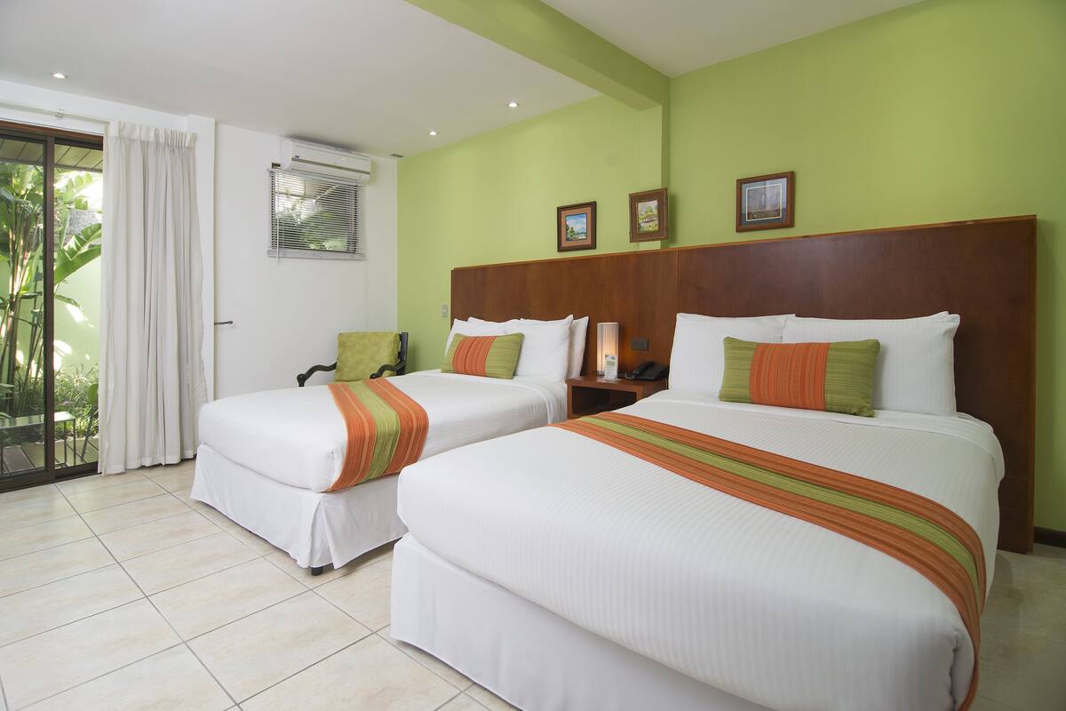 Habitación de hotel con dos camas, una puerta corrediza de vidrio a un balcón y una pared verde con cuadros enmarcados.