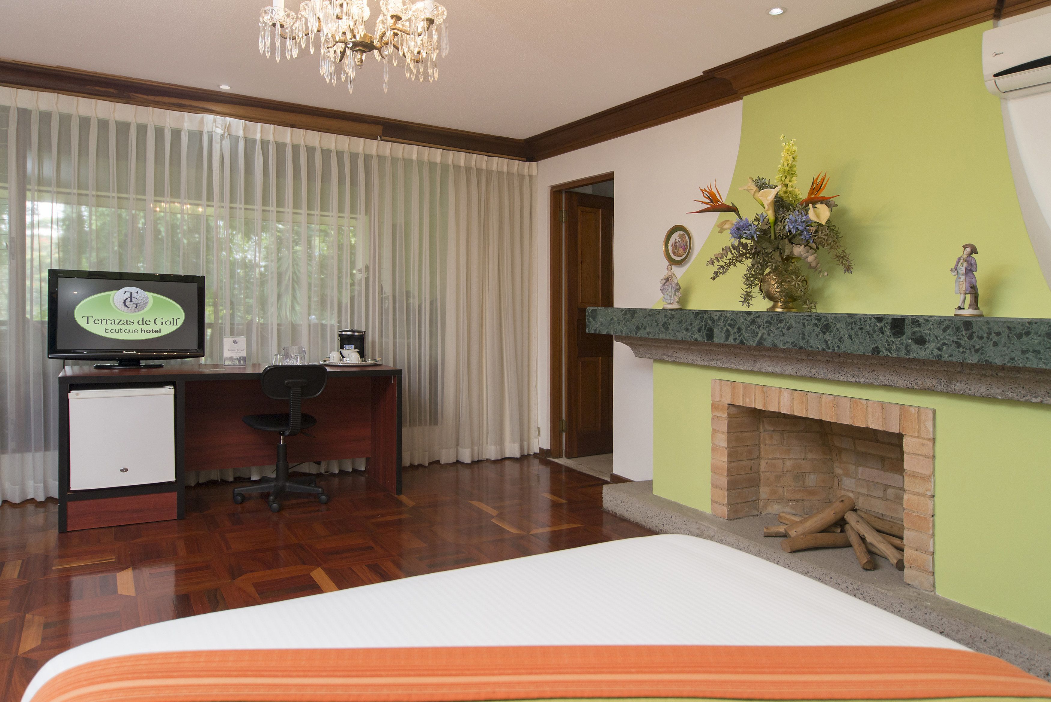 A hotel room with a bed, a desk with a computer, a television, a fireplace, and a window with curtains.