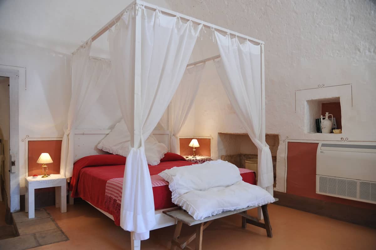 A four-poster canopy bed with white bedding, two bedside tables with lamps, and a small shelf with a decorative item.