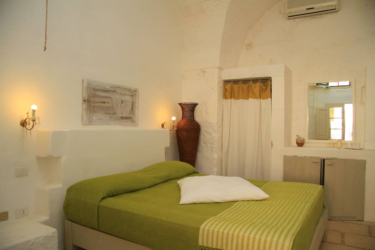 A bedroom with a bed, a large vase, a painting, and a mirror.