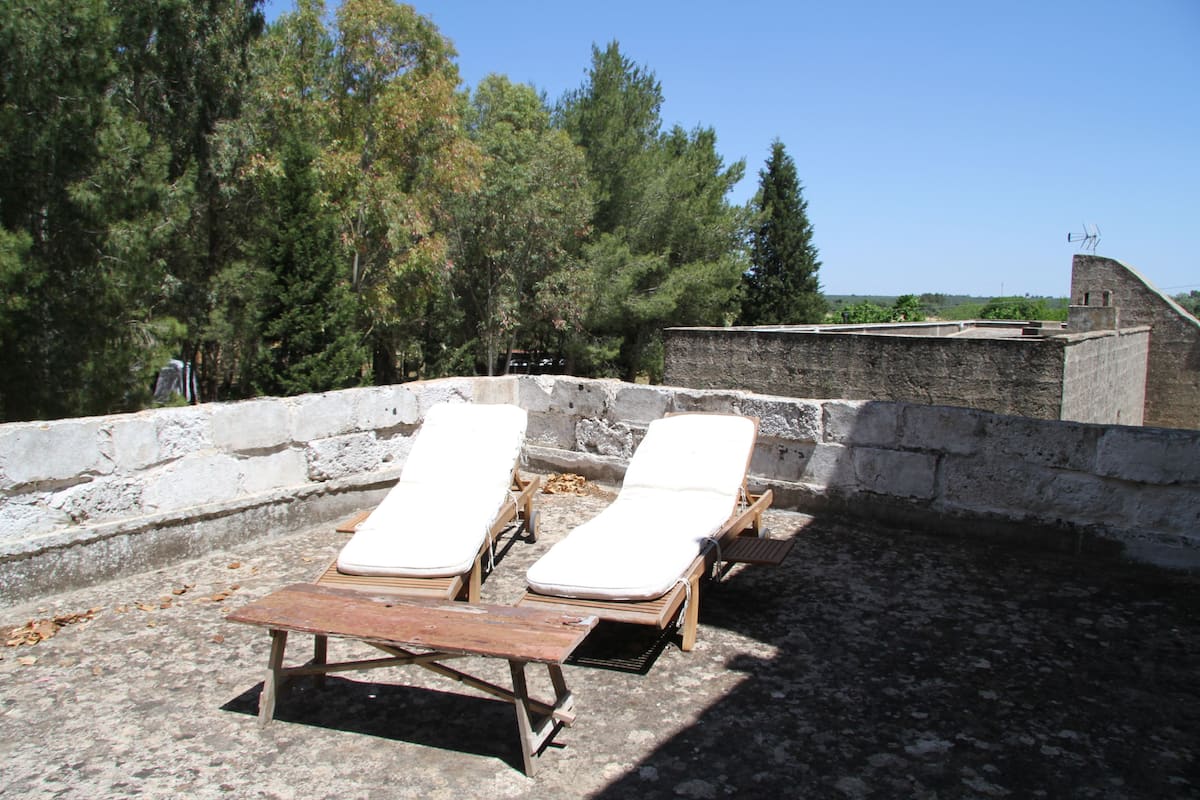 A rooftop terrace with two wooden lounge chairs and a table, surrounded by a stone wall and trees.