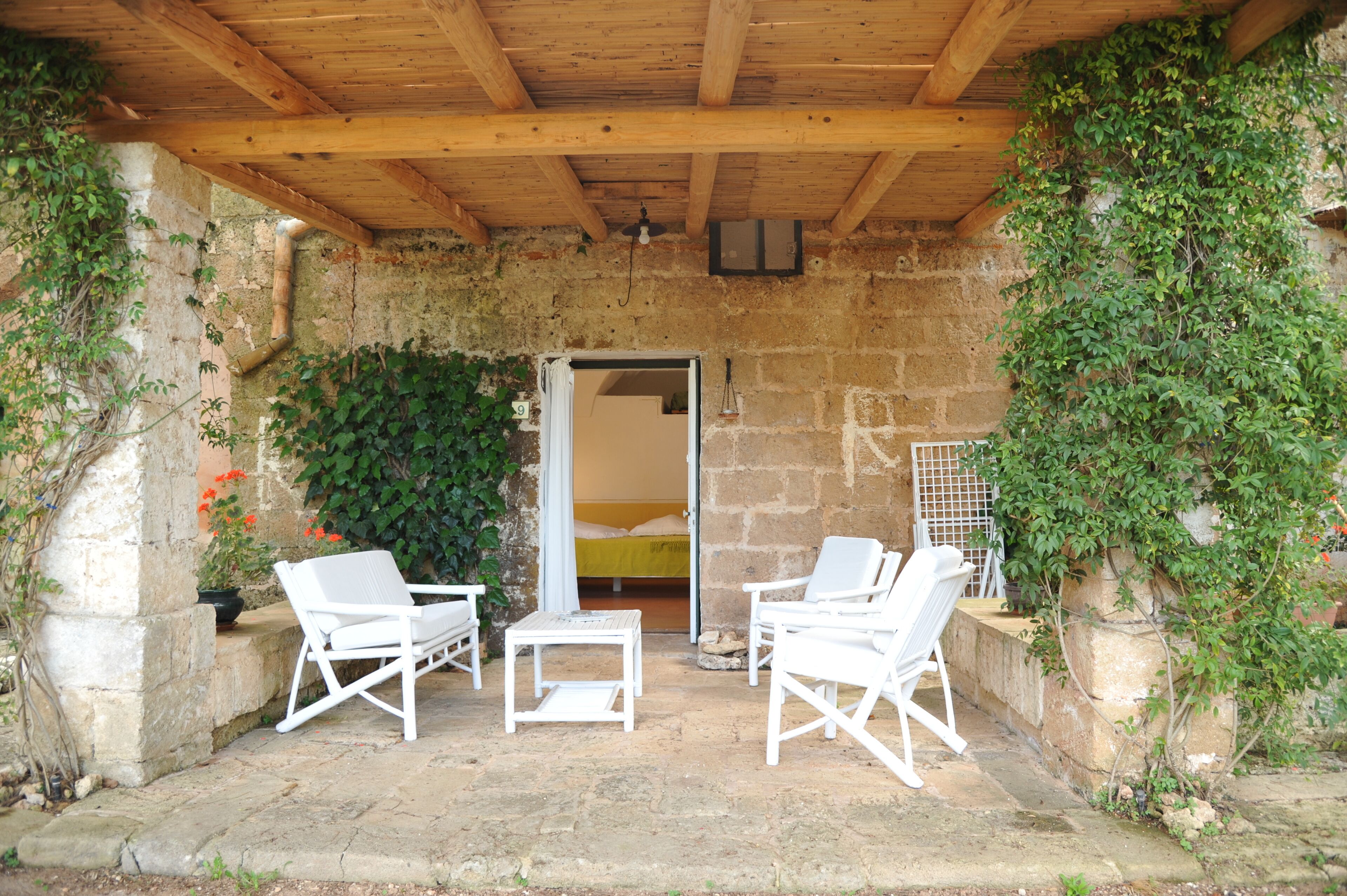 A covered outdoor seating area with white chairs and a table, a stone wall with greenery, and a doorway leading to a room.