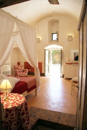 A bedroom with a canopy bed, a bedside table with a lamp, a chair, and a view of the outside. - Masseria Cristo (Ugento)