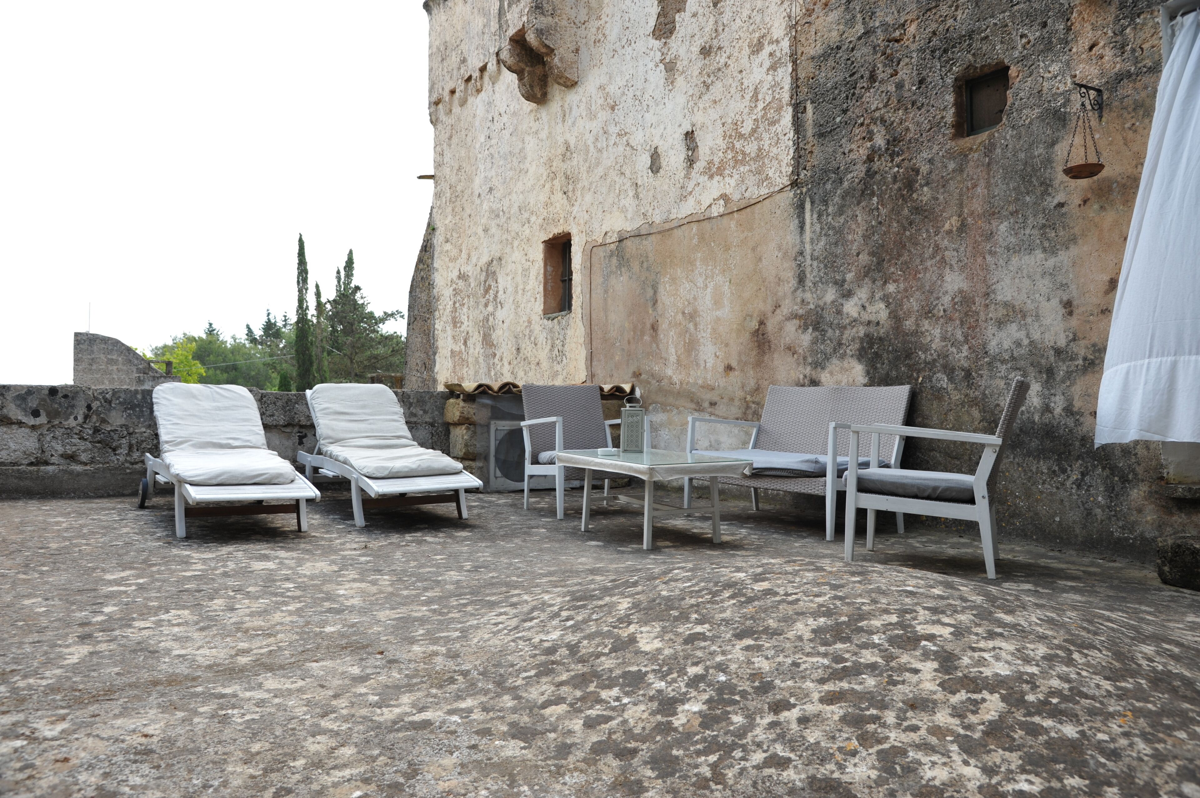 Eine Dachterrasse mit weißen Liegestühlen und einem Tisch, vor einer verwitterten Steinmauer.