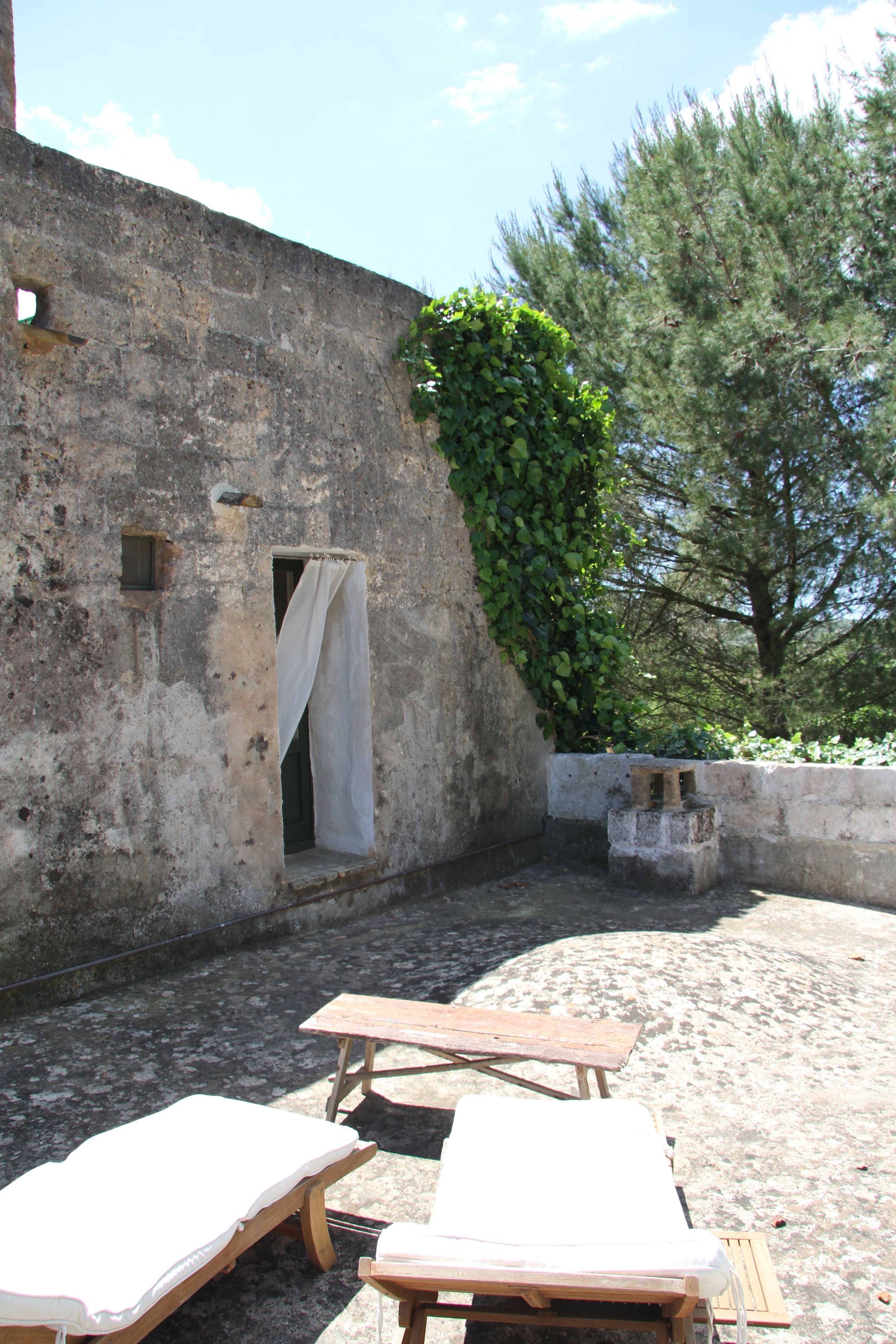A courtyard with a stone wall, a small window with a curtain, and two wooden lounge chairs.