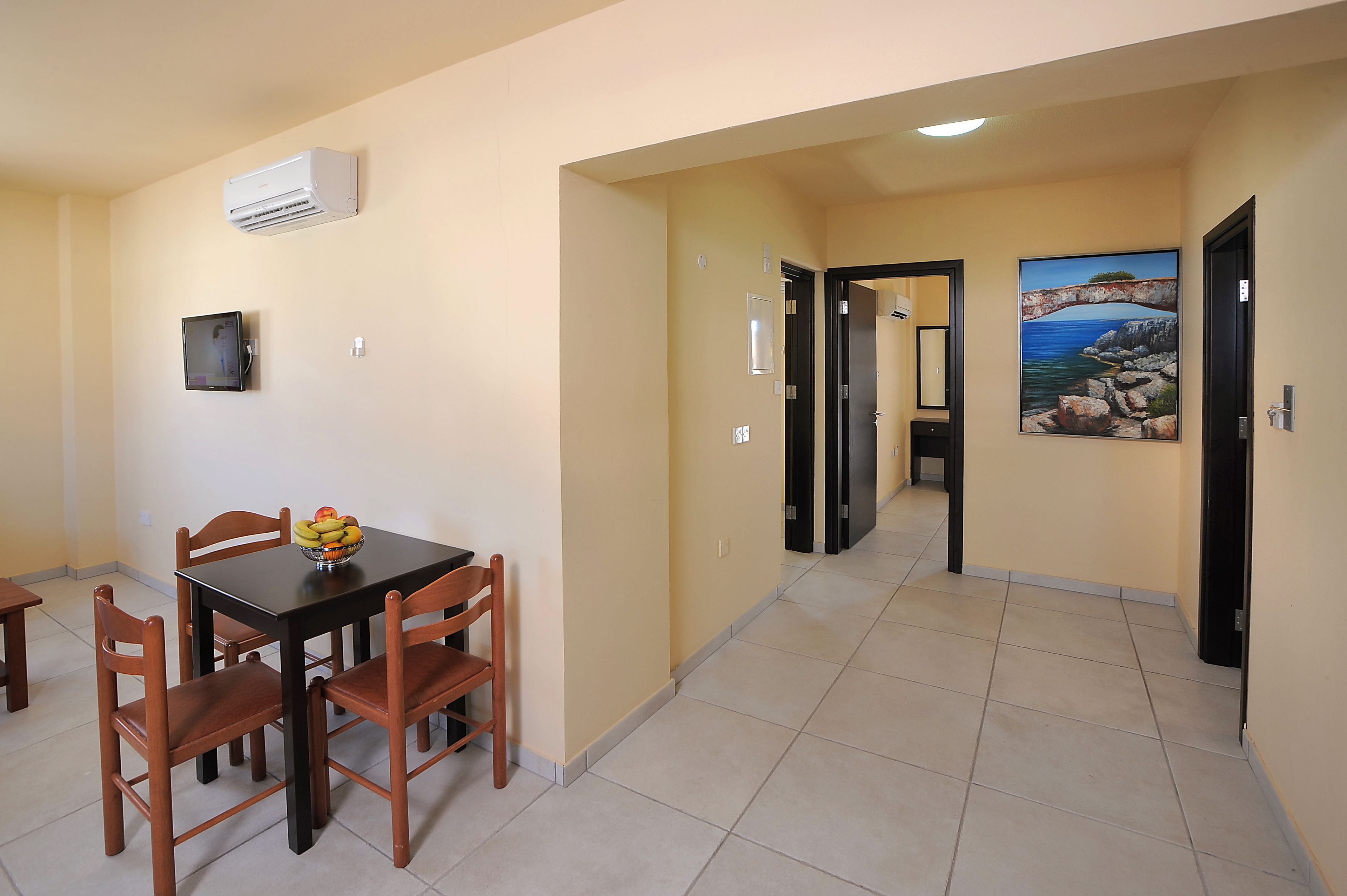 A dining area with a table and chairs, a wall-mounted air conditioner, a small television, and a painting of a coastal scene.