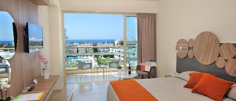 A hotel room with a large bed, a desk with a flat-screen TV, a balcony with a view of the city and ocean, and a modern decor style.