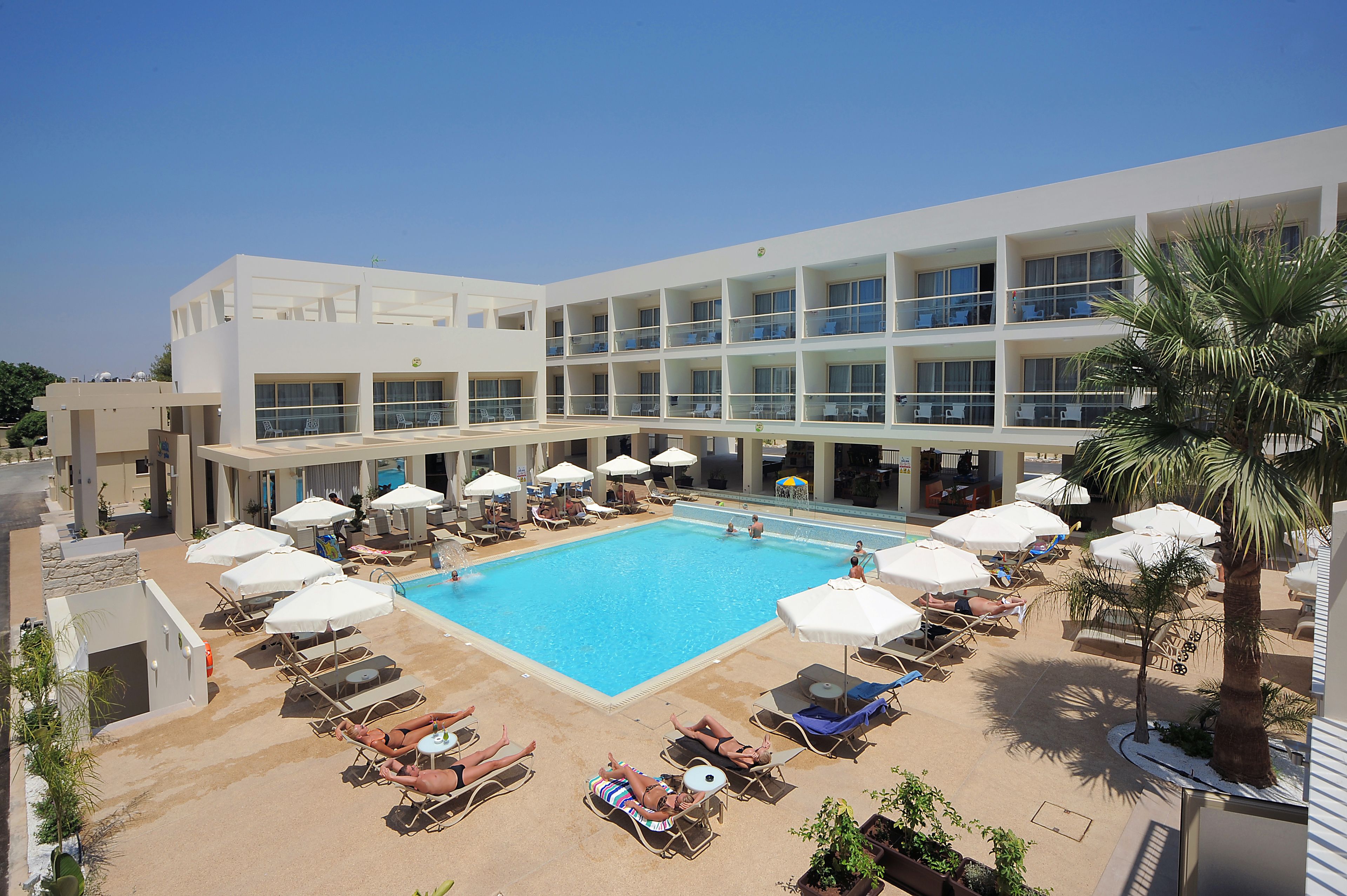A hotel with a large swimming pool surrounded by sun loungers and umbrellas, multiple balconies, and a palm tree.