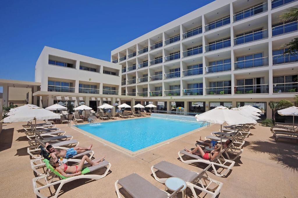 Un hotel con una gran piscina rodeada de tumbonas y sombrillas, un edificio de varios pisos con balcones, y un cielo azul despejado.