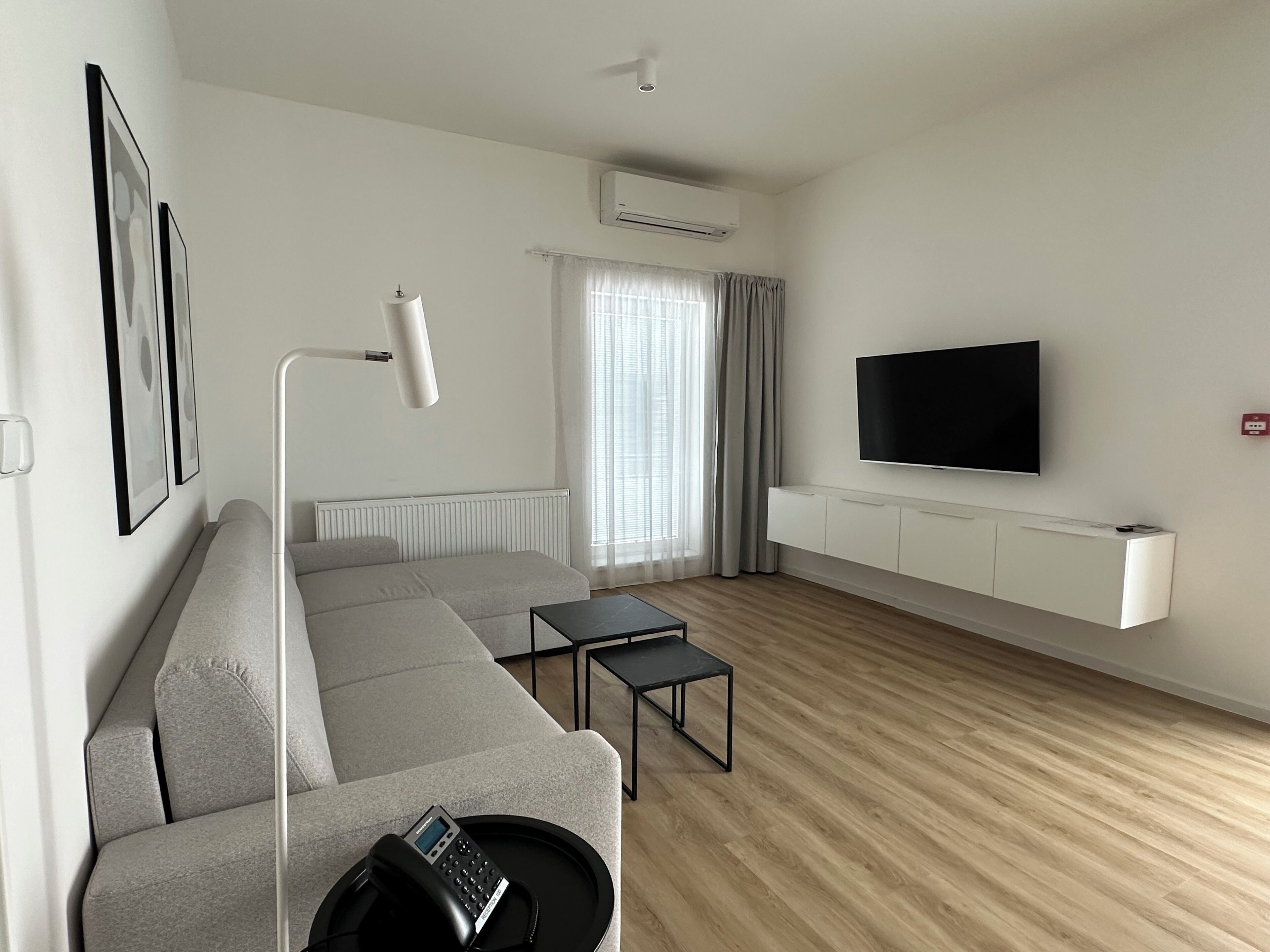 A modern living room with a grey sectional sofa, a small black table, a mounted TV, and an air conditioning unit on the wall.