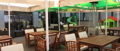 An outdoor patio area with wooden tables and chairs, umbrellas, and a green play structure.