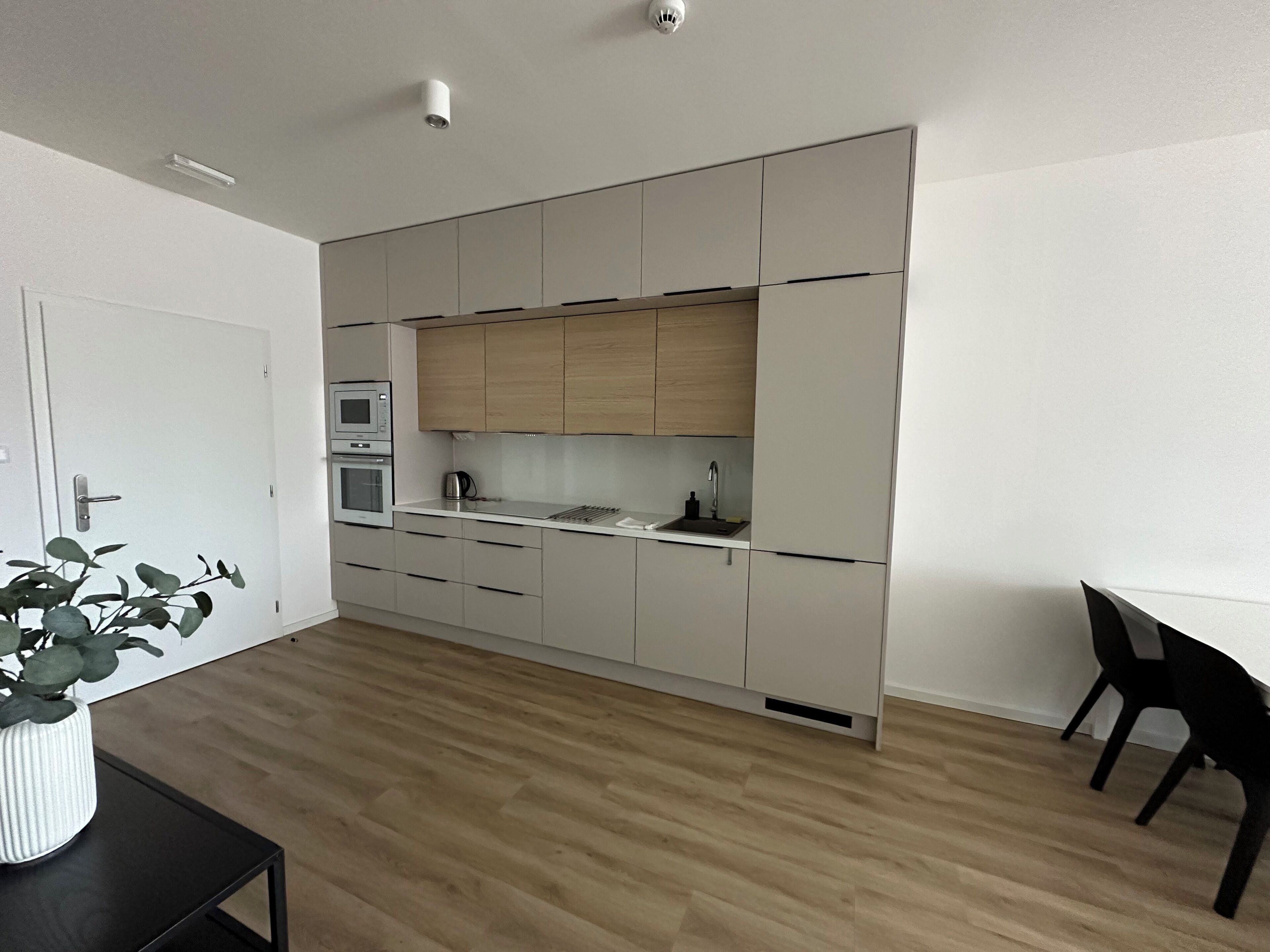 Et moderne køkken med hvide skabe, indbygget ovn, mikroovn og vask. Der er et spiseområde med et bord og stole, og en potteplante på et lille bord.