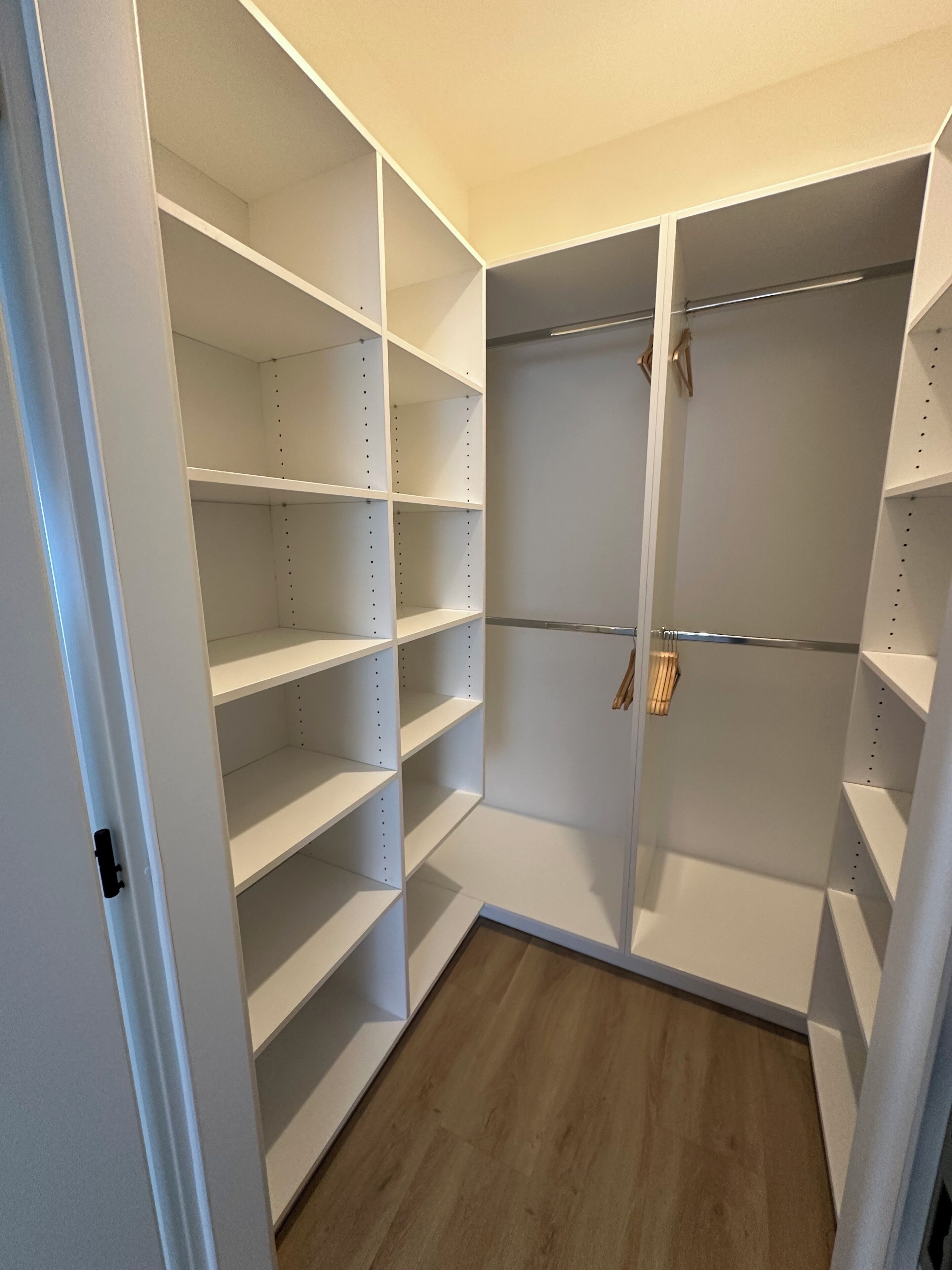 A walk-in closet with multiple shelves and hanging space, featuring a wooden floor and white walls.