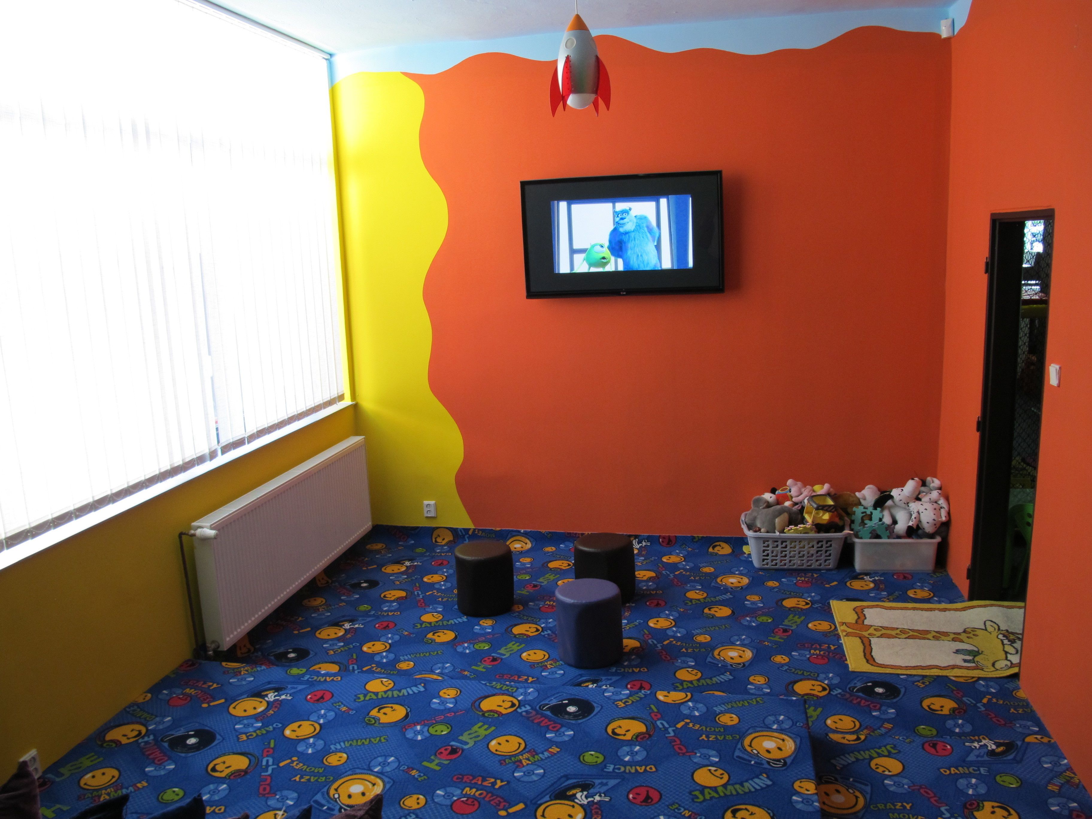 A room with a vibrant yellow and orange wall, a television mounted on the wall, a window with blinds, a radiator, a blue floor with smiley face patterns, and a basket of stuffed toys.