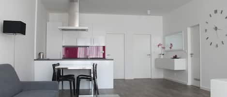 A modern living room with a grey sofa, a glass coffee table, and a kitchen area with white cabinets and a red backsplash.