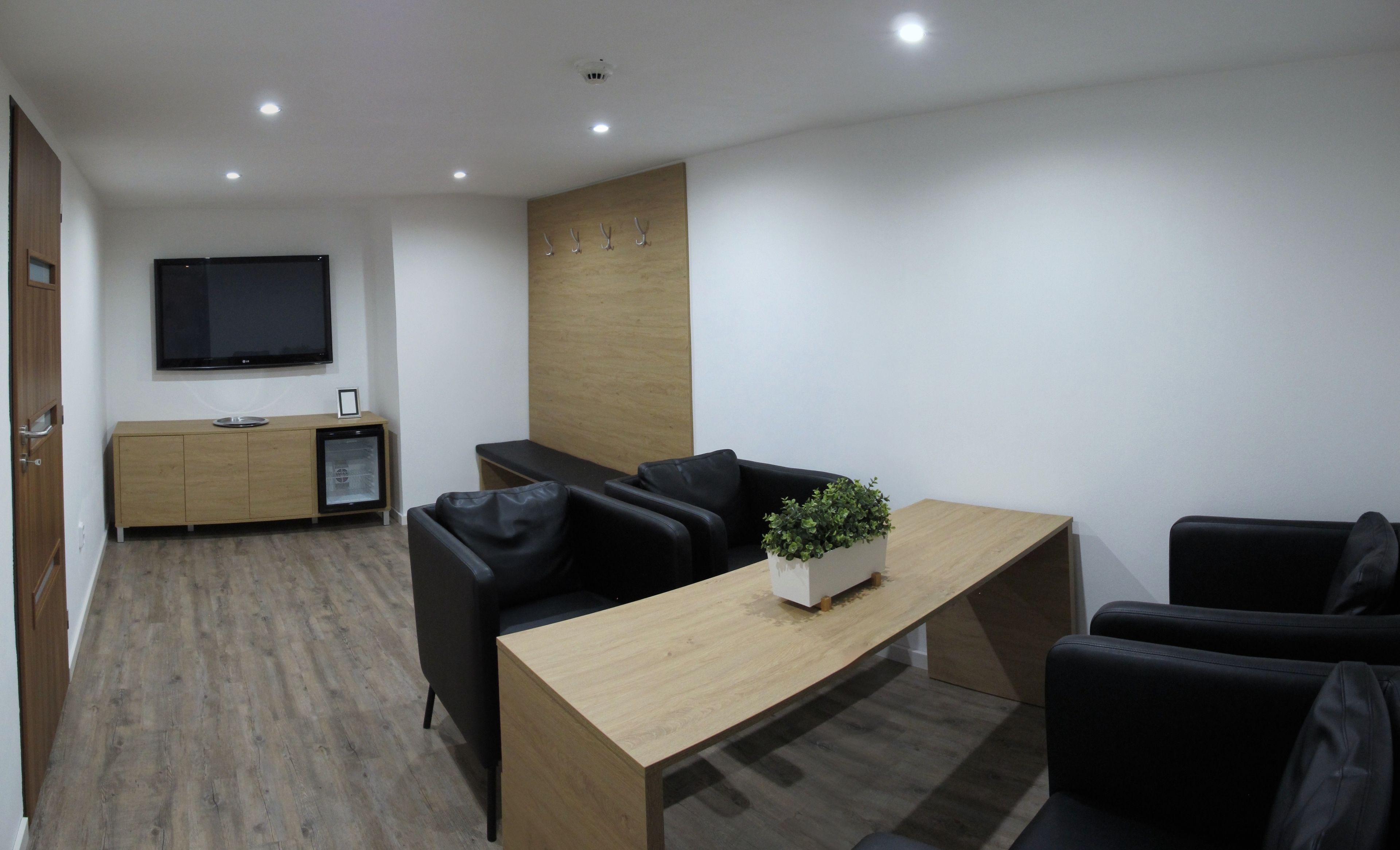 A modern living room with a wooden coffee table, black leather sofas, a flat-screen TV, and a small refrigerator.