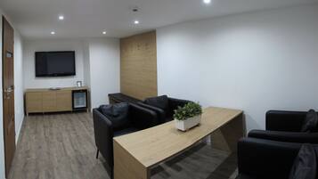 A modern living room with a wooden coffee table, black leather sofas, a flat-screen TV, and a small refrigerator.