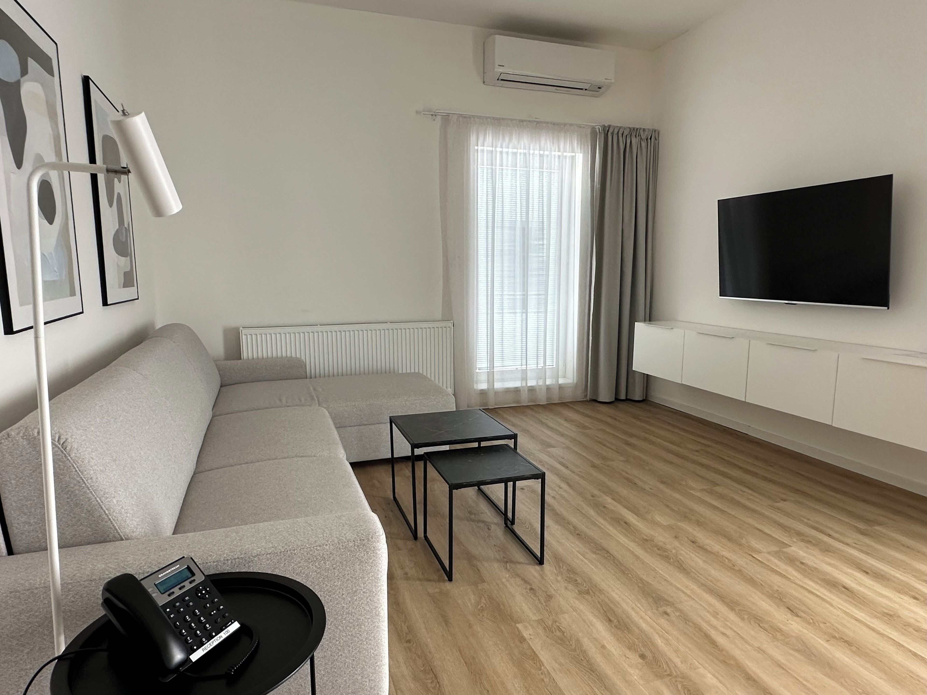 A modern living room with a grey sofa, a small black table, a mounted TV, and an air conditioning unit.