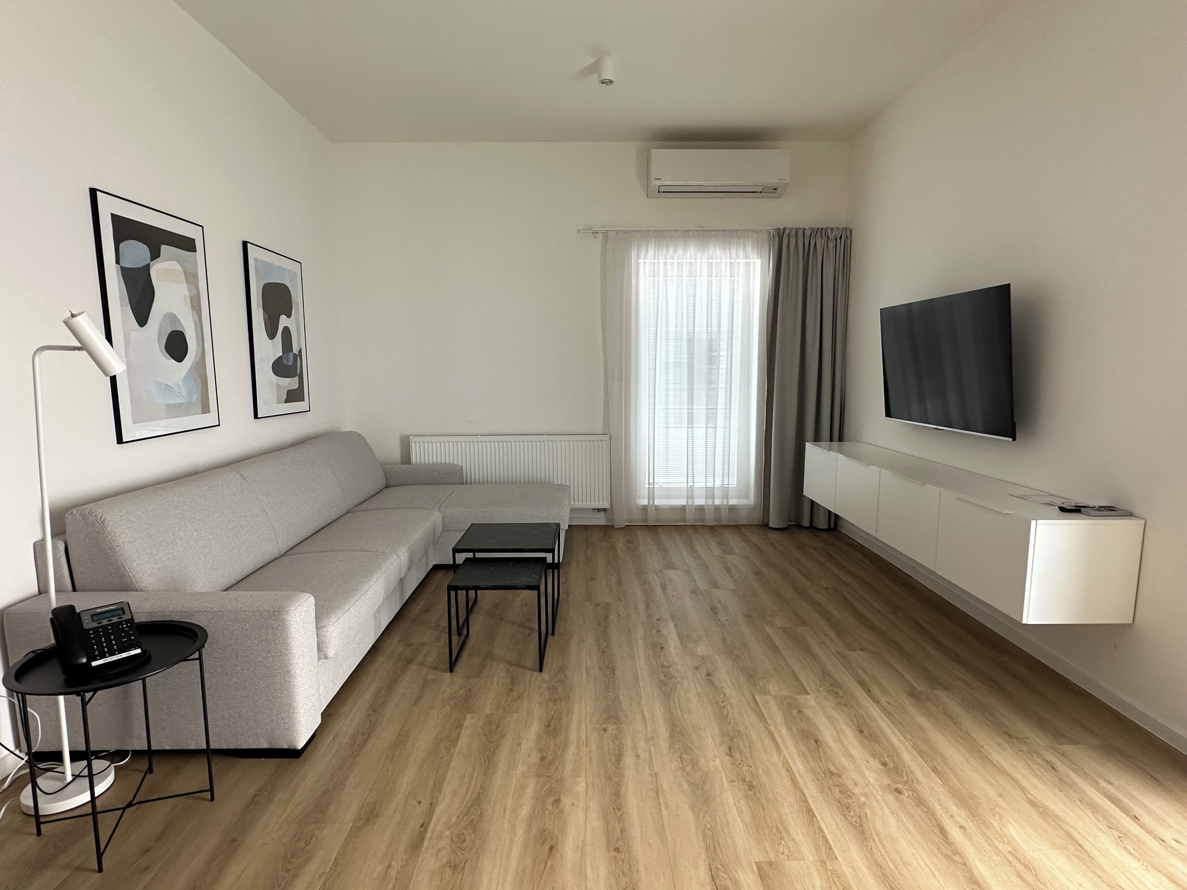 Una sala moderna con un sofá gris, una mesita de centro pequeña, una lámpara de pie, un televisor montado en la pared y un radiador.