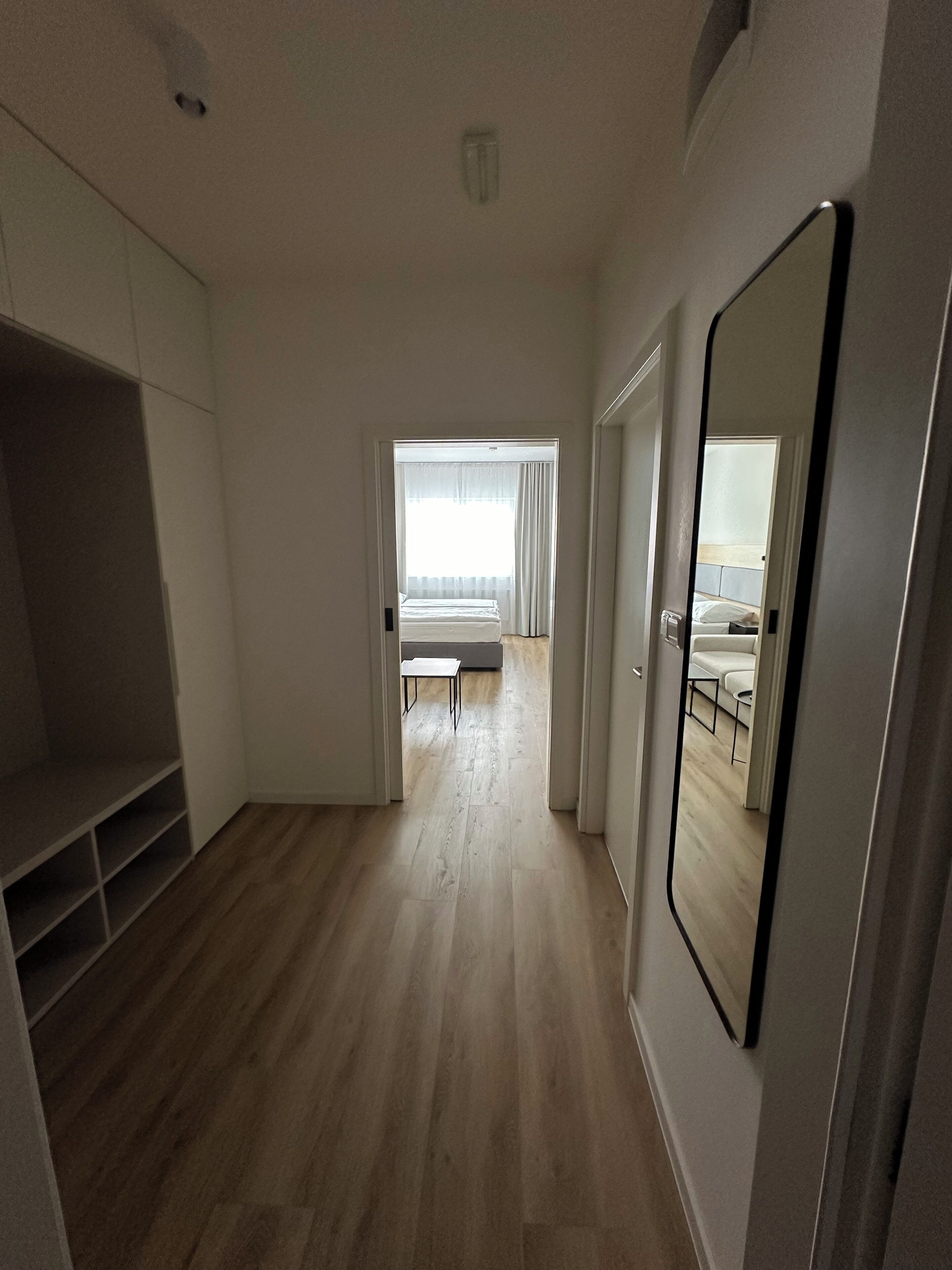 A modern, well-lit room with a wooden floor, a large mirror, and a view into a bedroom with a bed and curtains.