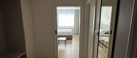 A modern, well-lit room with a wooden floor, a large mirror, and a view into a bedroom with a bed and curtains.