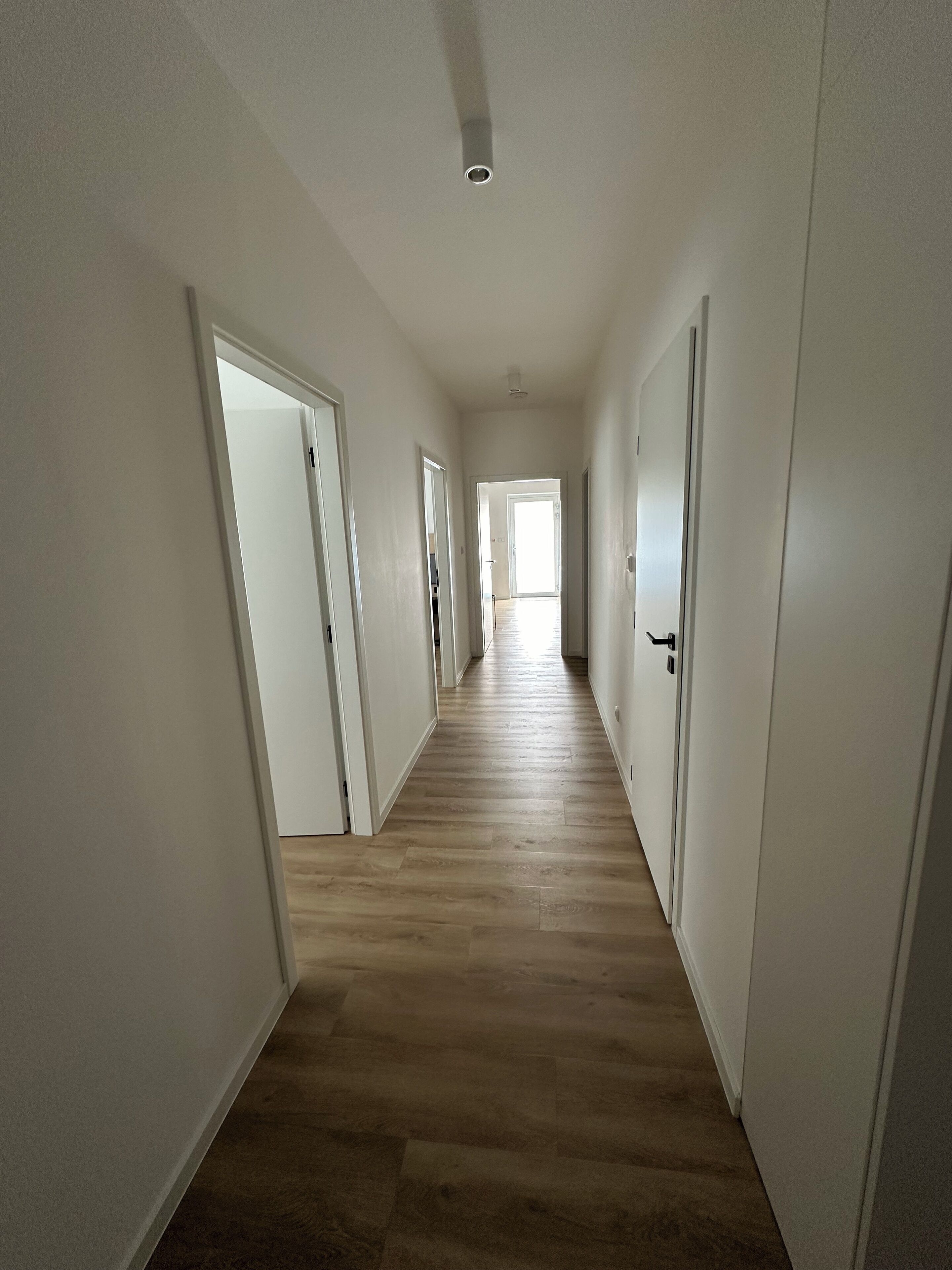 Un pasillo largo y estrecho con paredes blancas y suelo de madera, que conduce a una habitación luminosa al final.