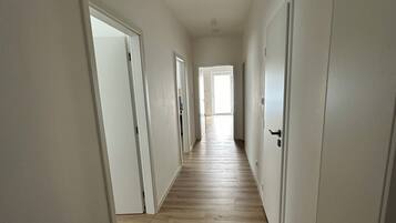 Un pasillo largo y estrecho con paredes blancas y suelo de madera, que conduce a una habitación luminosa al final.