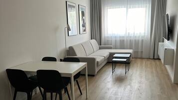 A modern living room with a sofa, a dining table with chairs, a TV, and a large window with curtains.