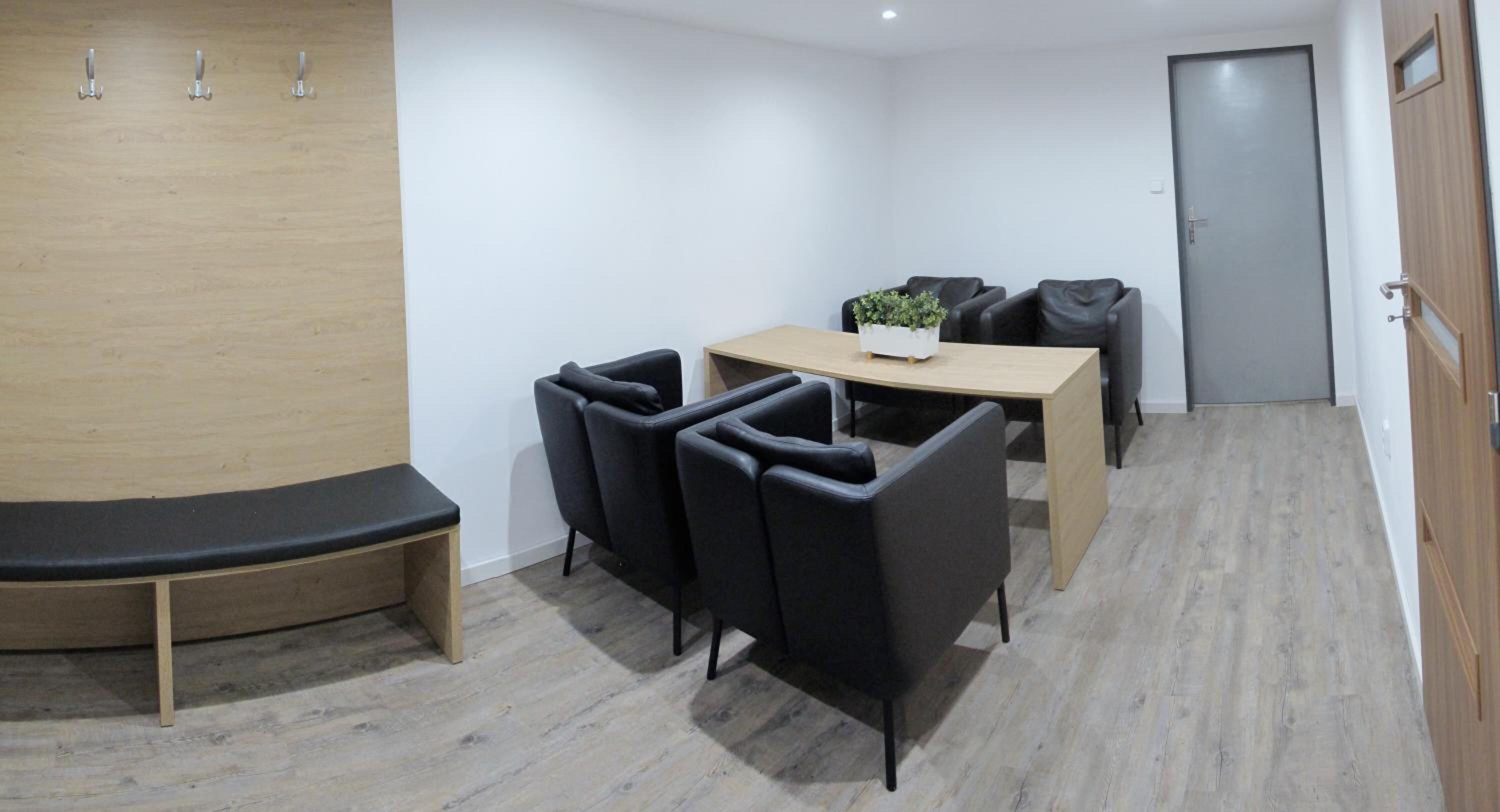 Une petite salle de réunion avec une table en bois, des chaises noires, une plante en pot et une porte au fond.