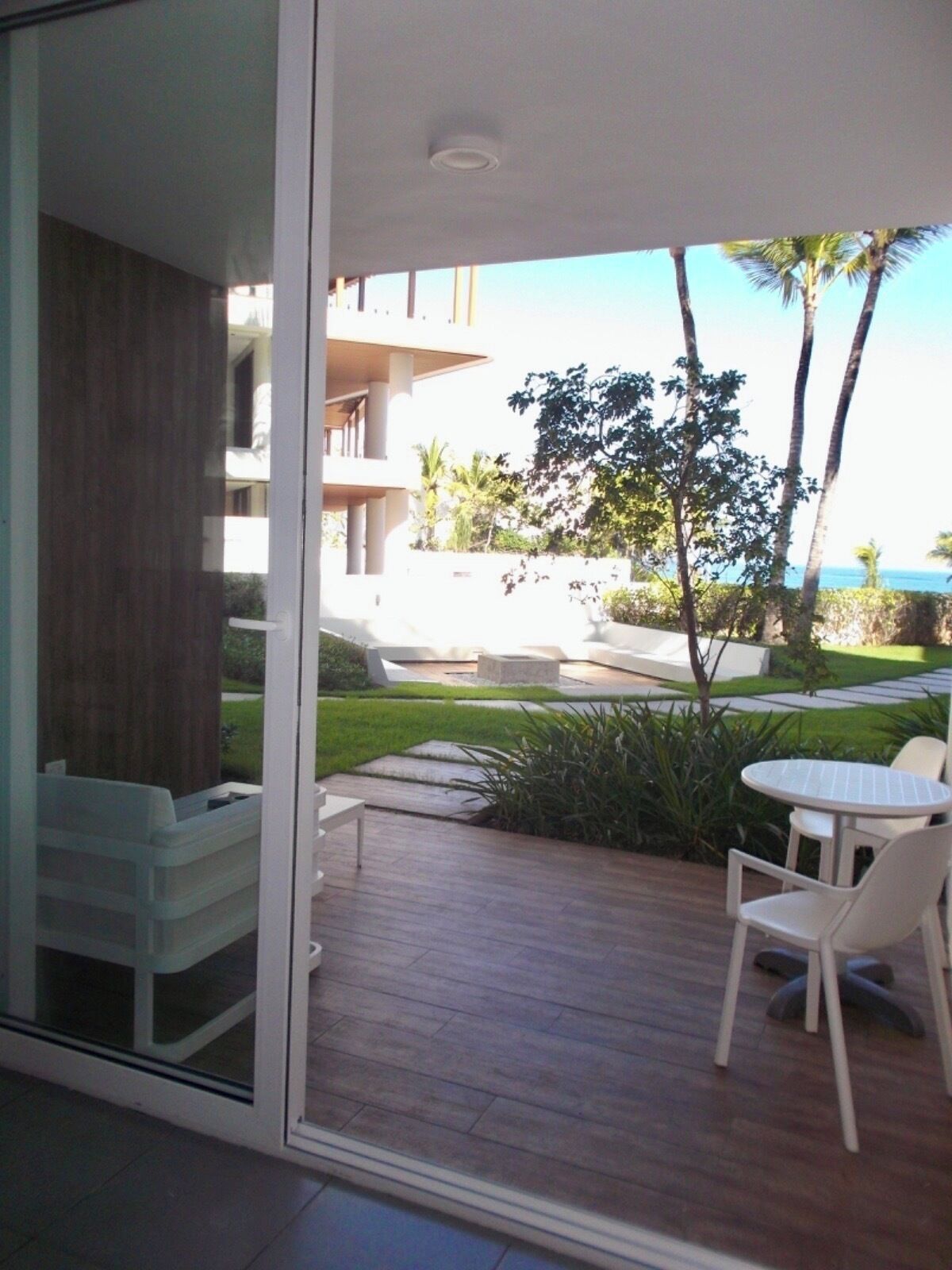 A modern outdoor patio with a white table and chairs, a small garden area, and a view of a building and ocean.