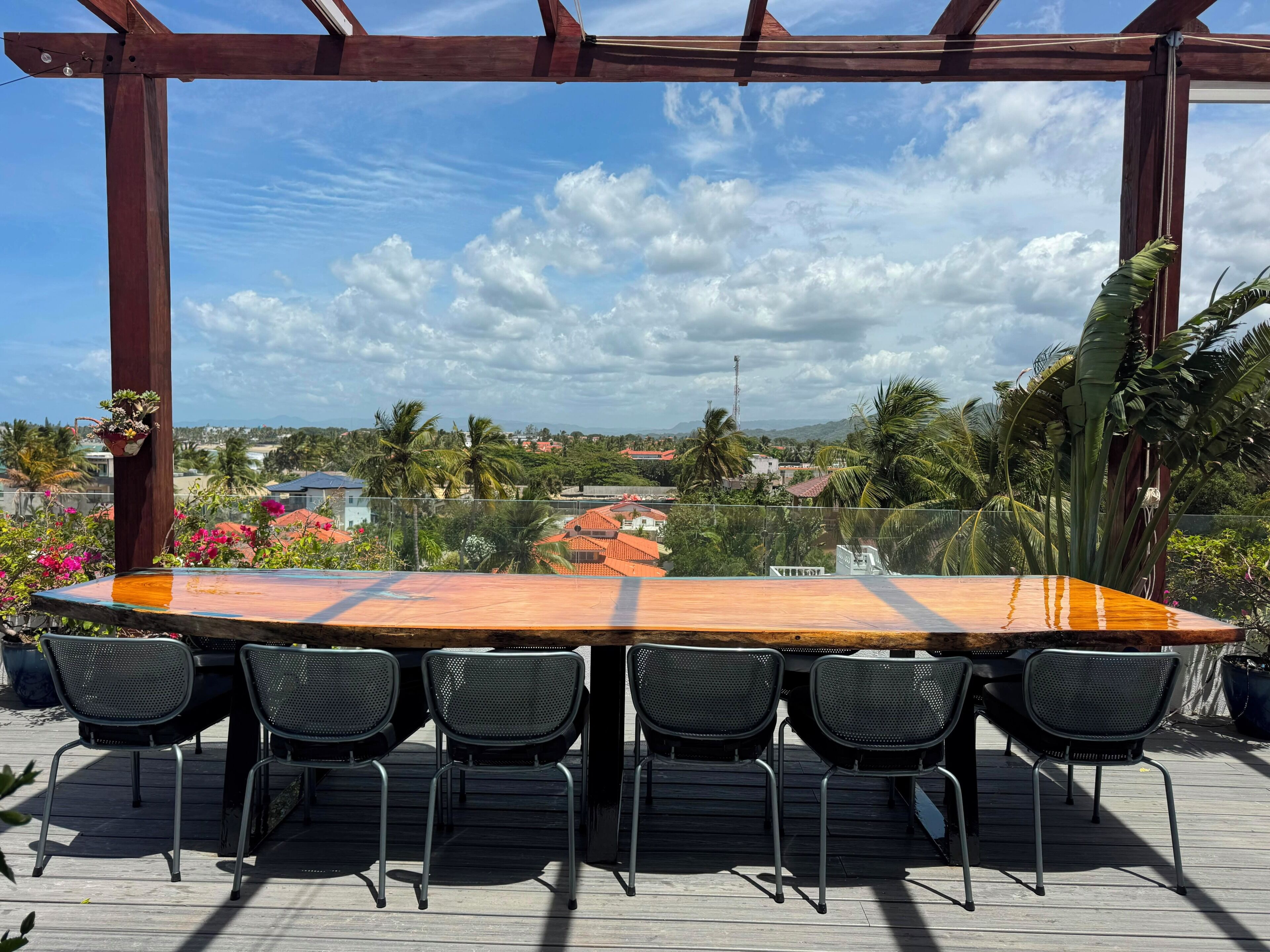 A wooden pergola with a table and chairs, overlooking a residential area with houses and palm trees.