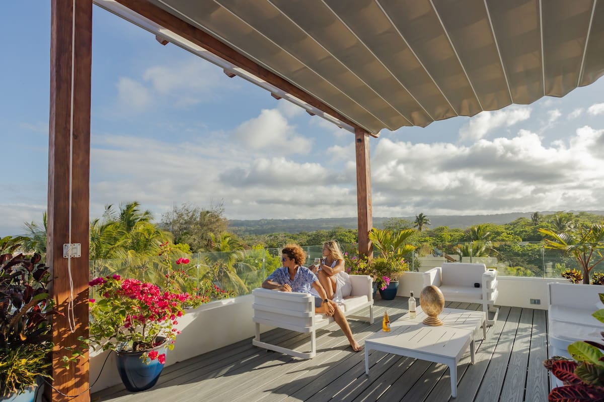 A covered outdoor seating area with white furniture, a wooden deck, and potted plants.
