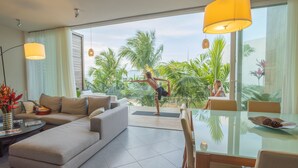 A modern living room with a large sofa, a glass dining table, and a view of palm trees and a body of water.