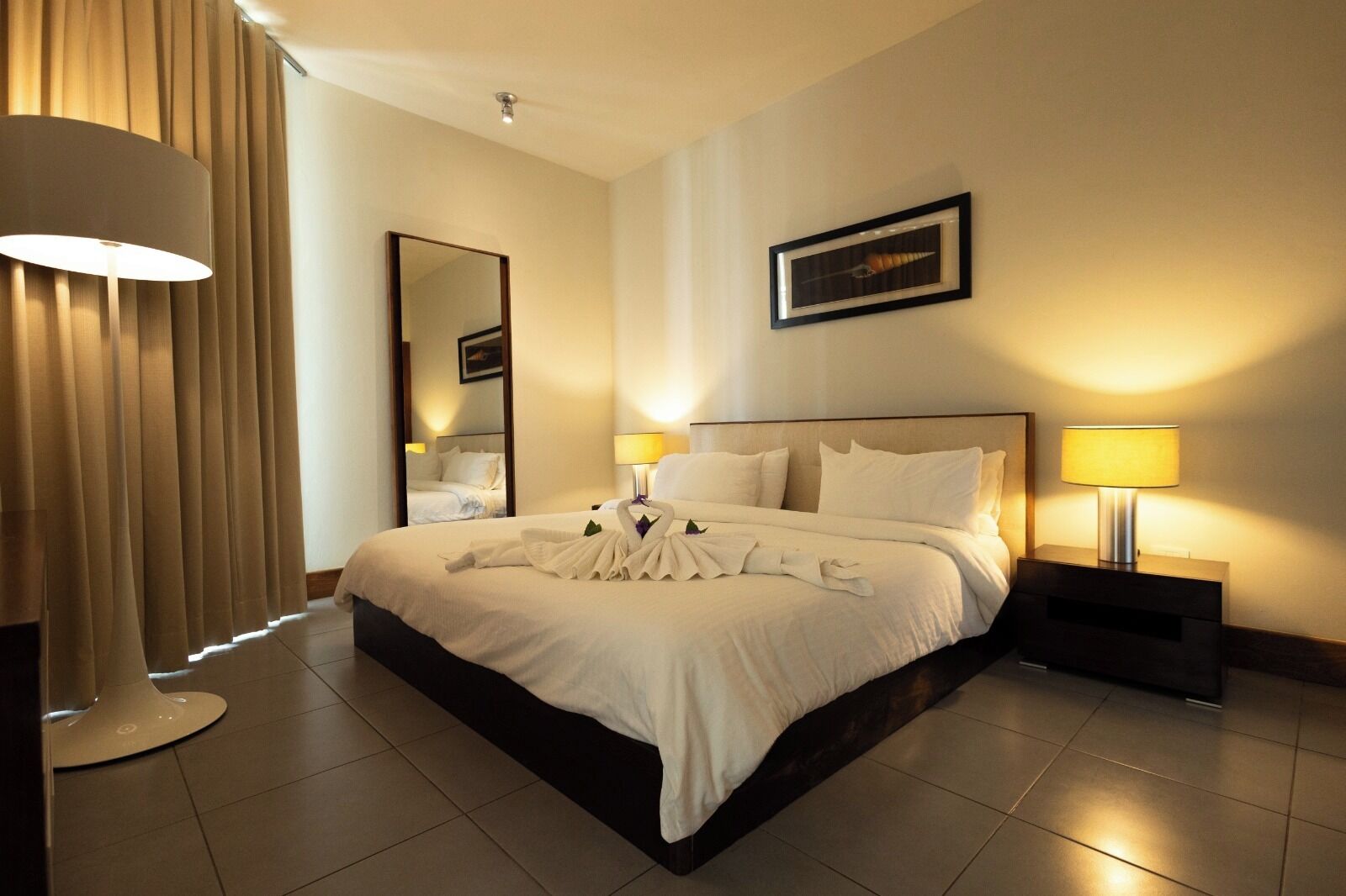 A hotel room with a large bed, two bedside lamps, a mirror, and a framed picture on the wall.