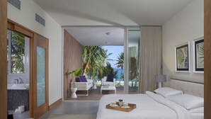 A modern hotel room with a large bed, a balcony with a view of palm trees and the ocean, a sitting area with a coffee table, and a bathroom with a large window. - Ultravioleta Boutique Residences (Cabarete)