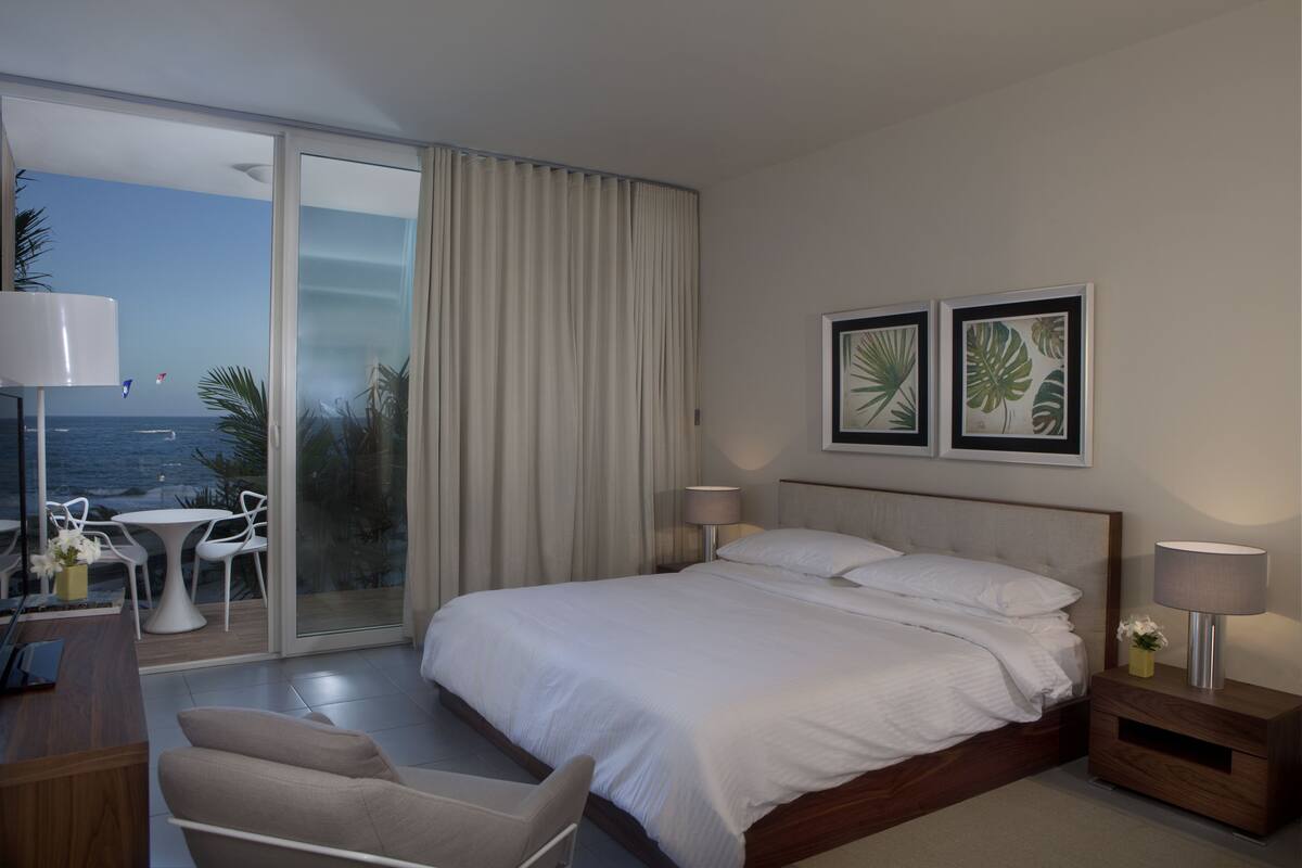 A modern hotel room with a large bed, a desk with a chair, a small table with chairs, and a balcony with a view of the sea.