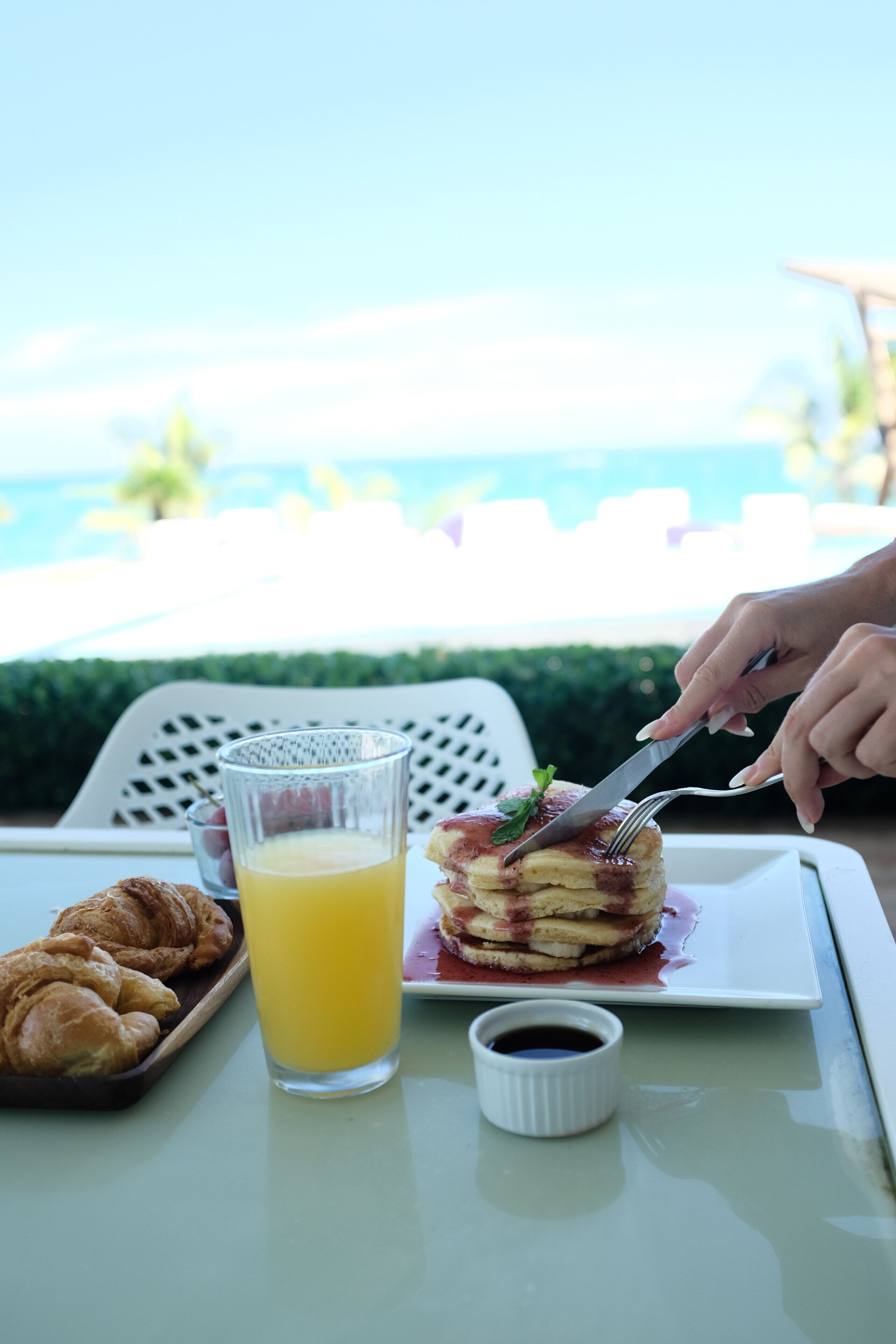 Un desayuno con cruasanes, un vaso de jugo de naranja y una pila de panqueques con mermelada y una hoja de menta sobre un plato blanco, con vista a una piscina al fondo.