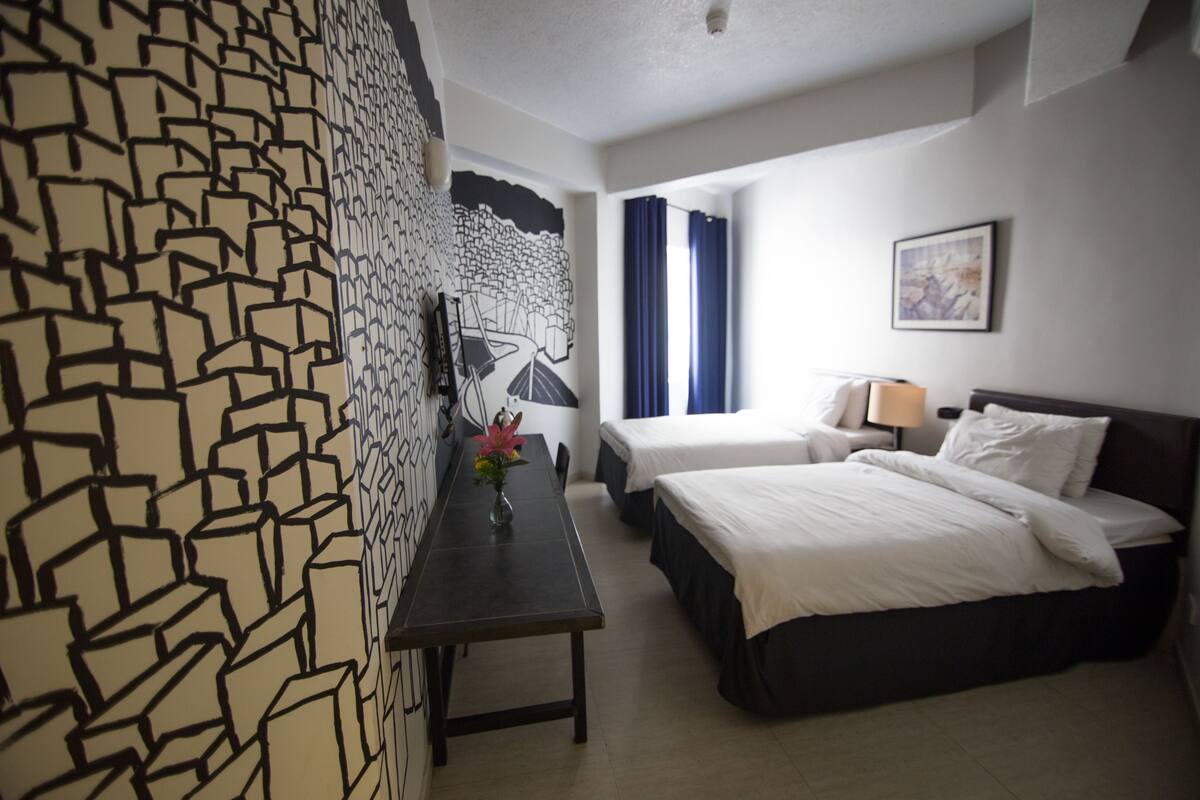 A hotel room with a patterned wall, a bed, a bedside table with a vase of flowers, a desk, and a chair.
